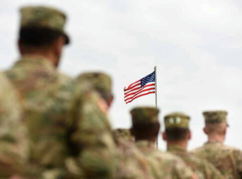 Soldiers in uniform standing, American flag in background.