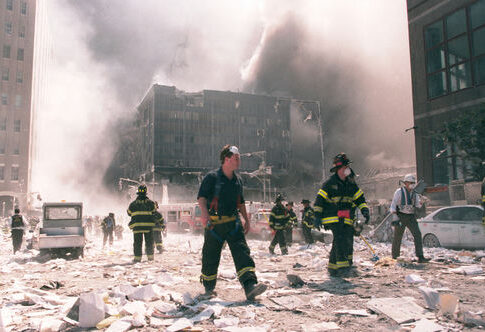 Firefighters walking through debris near a heavily damaged smoking building