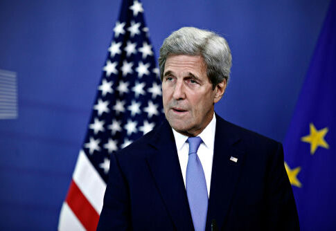 man in suit speaking at a podium with US and EU flags behind